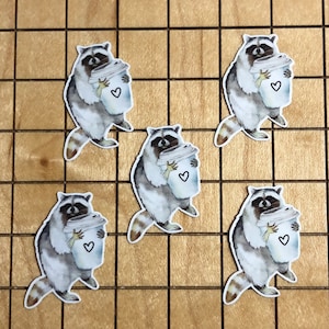 May include: Five watercolor stickers of raccoons holding coffee cups with hearts on them. The stickers are on a wooden background.
