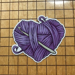 May include: A purple yarn ball in the shape of a heart with a crochet needle going through it.