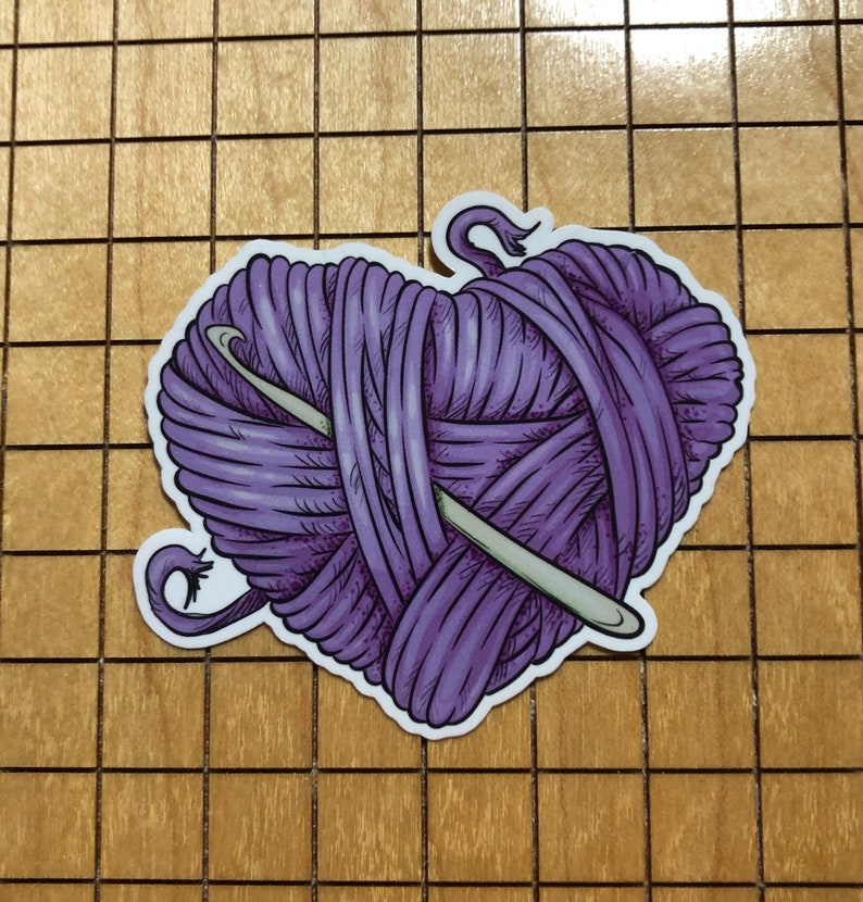 May include: A purple yarn ball in the shape of a heart with a crochet needle going through it.