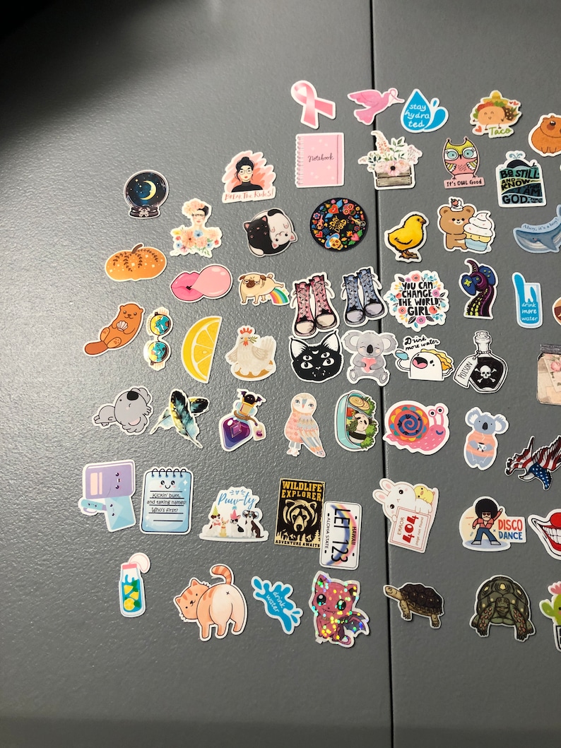 May include: A collection of colorful stickers featuring various designs, including animals, plants, food, and text. The stickers are arranged on a gray surface.