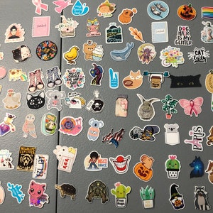 May include: A collection of colorful stickers featuring various designs, including animals, plants, text, and abstract patterns. The stickers are arranged on a gray surface.