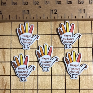 May include: Five Thanksgiving-themed hand-shaped embellishments. Each features a turkey design with a hand-shaped body and the text "Happy THANKS GIVING". The turkey's feathers are colored in shades of brown, orange, and yellow.