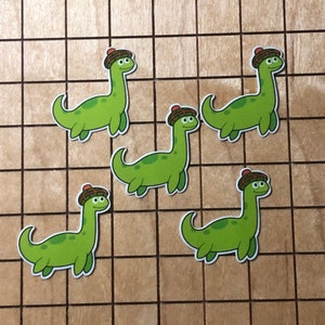 May include: Five green dinosaur stickers with brown and red hats. The dinosaurs are all facing forward with their mouths open.