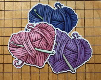 Könnte beinhalten: Drei herzförmige Wollknäuel mit Stricknadeln. Die Wollknäuel sind rosa, lila und blau.