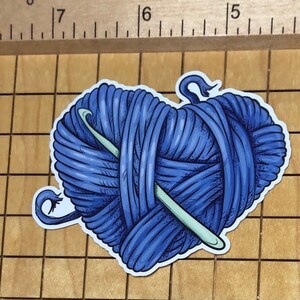 May include: A blue yarn ball with a crochet hook wrapped around it. The yarn ball is in the shape of a heart.
