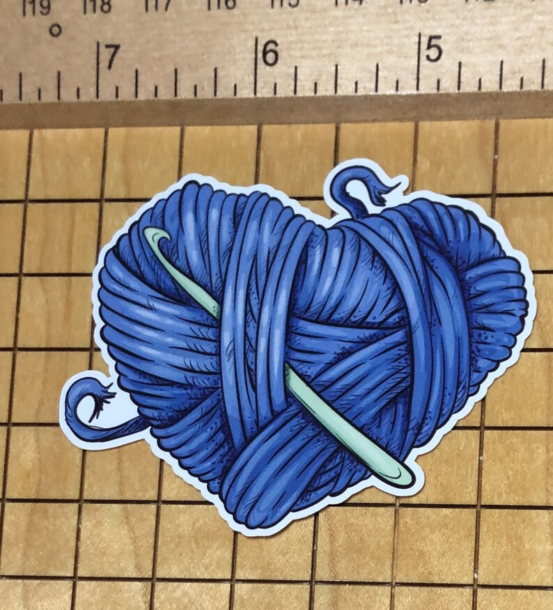 May include: A blue yarn ball with a crochet hook wrapped around it. The yarn ball is in the shape of a heart.