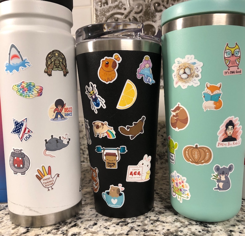 May include: Three stainless steel travel mugs with various colorful stickers. The mugs are black, white, and light blue. The stickers feature animals, food, and other designs. The text on the stickers includes "USA", "Disco Dance", "Happy Thanks Giving", "Error 404", "It's OWL Good", and "Break the Rules!"