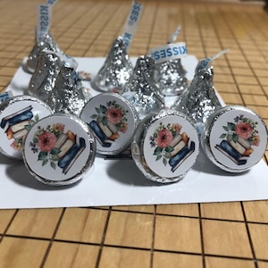 May include: A selection of foil-wrapped chocolate sweets, some with the word "Kisses" on the paper strip. Several sweets have a circular sticker with a floral and book design. The sweets are arranged on a white surface.