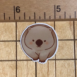 Puede incluir: Una pegatina de dibujos animados marr&oacute;n y blanca de un wombat con una expresi&oacute;n linda. El wombat est&aacute; enroscado en una bola con sus patas metidas debajo de su barbilla.