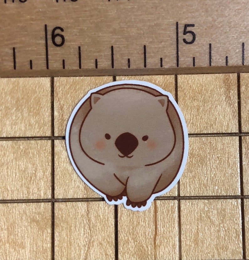 Puede incluir: Una pegatina de dibujos animados marr&oacute;n y blanca de un wombat con una expresi&oacute;n linda. El wombat est&aacute; enroscado en una bola con sus patas metidas debajo de su barbilla.