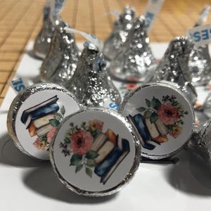 May include: Close-up of Hershey's Kisses candies wrapped in silver foil. The candies have round stickers with a watercolor design of books and flowers. The stickers feature a stack of books with a floral arrangement of pink and yellow flowers.