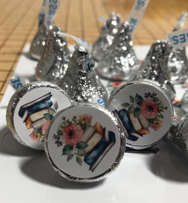 May include: Close-up of Hershey's Kisses candies wrapped in silver foil. The candies have round stickers with a watercolor design of books and flowers. The stickers feature a stack of books with a floral arrangement of pink and yellow flowers.