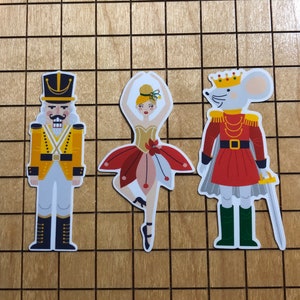 May include: Three paper cutouts of characters from the Nutcracker: a red and gold soldier, a ballerina with a red and white tutu, and a mouse wearing a red and gold crown.