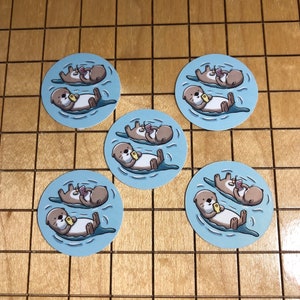 May include: Five circular stickers featuring a light blue background with two brown otters floating on their backs in water. One otter holds a yellow rectangular object, while the other holds a pink rectangular object. The design is repeated on all five.