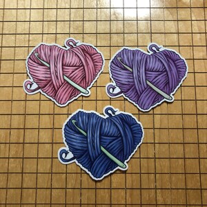 May include: Three heart-shaped yarn balls with a crochet hook through each. The yarn balls are pink, purple, and blue.