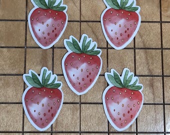 May include: Five strawberry stickers. Each sticker features a red strawberry with small seeds and a green leafy top. The stickers are on a light brown, grid-patterned surface.