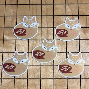 May include: Five brown and white fox stickers with a leaf design. The foxes are curled up and have closed eyes.