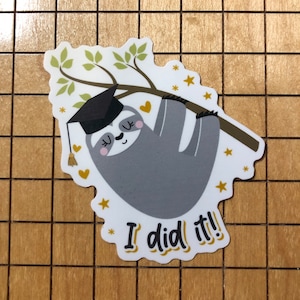 May include: A white sticker with a cartoon sloth wearing a graduation cap and hanging from a branch. The sloth is gray with a pink nose and black eyes. The sticker has gold stars and hearts around the sloth. The text "I did it!" is written in gold at the bottom of the sticker.