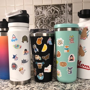 May include: Three stainless steel water bottles with various colorful stickers. The bottles are black, white, and light blue. The stickers feature animals, food, and other designs.