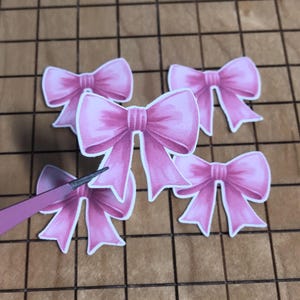 May include: Five pink paper bows with white outlines are arranged on a brown grid surface. A pair of pink tweezers is holding one of the bows. The bows have a watercolor effect.