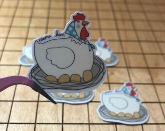 May include: A white chicken with a red comb and blue polka dot scarf sits on a nest of eggs. The text "mama hen" is written on the chicken's body.
