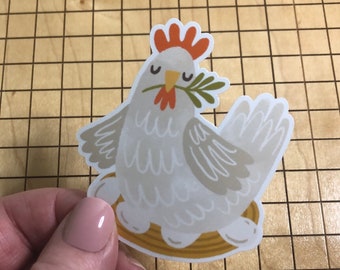 May include: A white chicken sticker with a red comb and a green sprig in its beak. The chicken is sitting on a yellow nest.