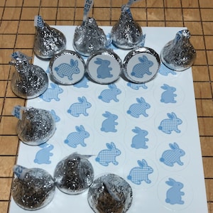 Könnte beinhalten: Eine Auswahl an folienverpackten Schokoladenbonbons und blauen Hasenaufklebern auf einer weißen Oberfläche. Die Aufkleber zeigen ein hellblaues Hasenmuster, einige mit einem Karomuster. Die Süßigkeiten haben Papieranhänger.