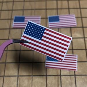 Könnte beinhalten: Kleine rechteckige amerikanische Flaggen mit roten und weißen Streifen und einem blauen Sternenfeld. Eine Flagge wird von einer rosa Pinzette gehalten, drei weitere liegen auf einer braunen, gefliesten Oberfläche. Es handelt sich um Aufkleber oder Dekorationen.