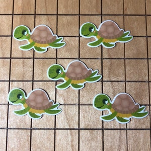May include: Five cartoon turtle stickers with brown shells and green bodies. The turtles have big eyes and are smiling. The stickers are on a wooden surface.