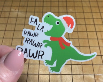 May include: A green dinosaur sticker wearing a Santa hat and red scarf. The sticker has the text "FA LA RAWR RAWR RAWR" in black font. The dinosaur is on a white background.