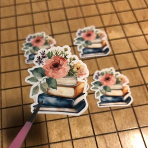 May include: Four stickers featuring stacks of books topped with floral arrangements. The books are in shades of blue and brown, with the flowers in pink, yellow, and green. The stickers are on a wooden surface.