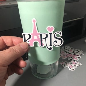 May include: A mint green tumbler with a sticker of the Eiffel Tower and the word "PARIS" in black with a pink heart.