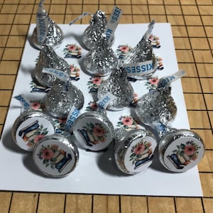 May include: A collection of Hershey's Kisses candies wrapped in silver foil. Some candies have a sticker with a floral design and books. The candies are arranged on a white card with a floral and book pattern.