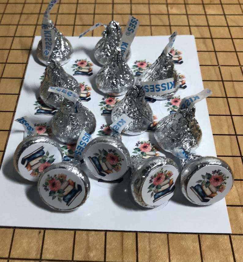 May include: A collection of Hershey's Kisses candies wrapped in silver foil. Some candies have a sticker with a floral design and books. The candies are arranged on a white card with a floral and book pattern.