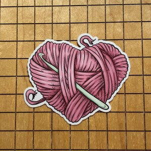 May include: A pink yarn ball in the shape of a heart with a knitting needle sticking out of it.