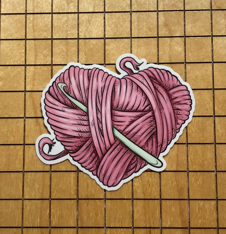 May include: A pink yarn ball in the shape of a heart with a knitting needle sticking out of it.