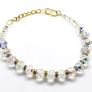 Op de afbeelding: Een delicate armband met heldere, iriserende kristallen kralen afgewisseld met goudkleurige metalen accenten. De armband heeft een goudkleurige ketting en sluiting, wat een sprankelend en elegant accessoire creëert. De kristallen reflecteren een regenboog aan kleuren.