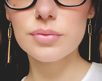 Peut inclure: Boucles d'oreilles en chaîne dorée avec un pendentif long et fin en forme de barre. Les boucles d'oreilles sont portées par une personne aux cheveux bruns et aux lunettes.