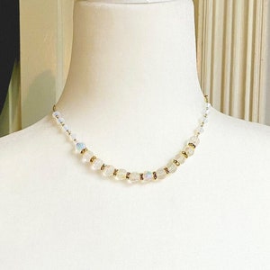Op de afbeelding: Een delicate witte en gouden kralenketting met een eenvoudig ontwerp. De ketting is gemaakt van kleine, ronde kralen die aan elkaar geregen zijn op een dunne ketting. De kralen zijn licht, iriserend wit met een subtiele gouden glans.