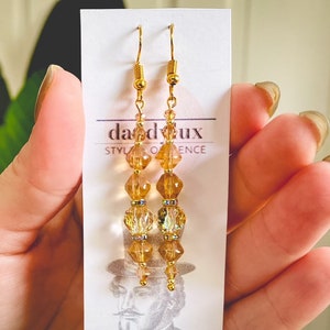 Op de afbeelding: Gouden oorhangers met hangende amberkleurige glazen kralen. De oorhangers bevinden zich op een witte kaart met de tekst "daddeux style & essence".