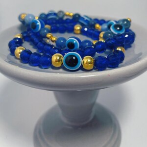May include: A stack of blue and gold beaded bracelets with evil eye charms. The bracelets are on a gray ceramic pedestal.