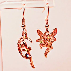 May include: A pair of gold-tone fairy dangle earrings. One earring features a fairy flying around a crescent moon, while the other earring features a fairy with outstretched wings.