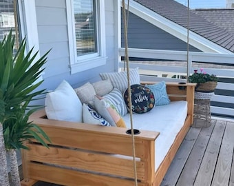 May include: A wooden swing bed with a white mattress and pillows. The swing bed is hanging from the ceiling of a porch with a white railing. There is a potted plant on the left side of the image and a small table with a wicker basket on the right side of the image. The pillow on the swing bed has the text "good vibes" on it.