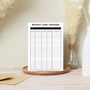 Może przedstawiać: Biała karta do śledzenia utraty wagi z czarnymi liniami i tekstem, wyświetlana na drewnianej tacy. Tekst u góry brzmi "WEIGHT LOSS TRACKER". Karta jest otoczona dekoracyjnymi wazonami i suszonymi roślinami.