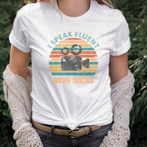 May include: White t-shirt with a vintage-style graphic of a film camera and the text "I Speak Fluent Movie Quotes".