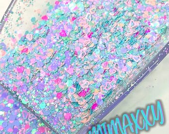 May include: A close-up of a clear container filled with iridescent glitter in various shades of blue, pink, and green. The glitter is shaped like hearts and stars. The text "minakku" is written in blue letters at the bottom of the image.