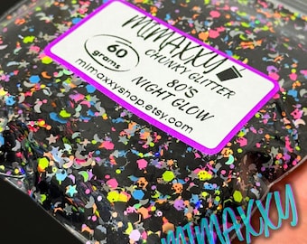 Può includere: Un sacchetto di plastica trasparente pieno di glitter grossi in varie forme e colori. I glitter sono principalmente neri con accenti rosa neon, gialli, blu e viola. L'etichetta recita "MIMAXXY CHUNKY GLITTER 80'S NIGHT GLOW" e contiene 60 grammi.