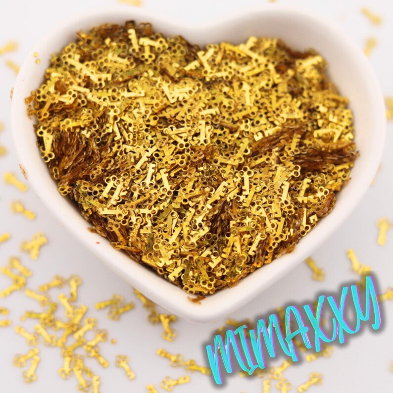 May include: A heart-shaped white bowl filled with small, gold-colored key-shaped embellishments. The keys are scattered throughout the bowl and some are outside the bowl. The text "MIMAXXXU" is written in a teal color at the bottom right.