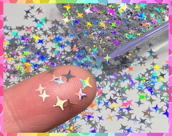 May include: A close-up of iridescent holographic star-shaped glitter. The glitter is scattered on a white surface and a finger is holding some of the glitter.