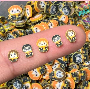 May include: A collection of polymer clay slices featuring cartoon characters in yellow, black, and orange. Several slices are on a fingertip, with many more scattered in the background. The characters have round faces and are wearing scarves and uniforms.
